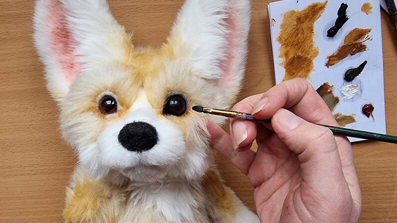 May include: A close-up of a plush toy fox with brown fur, white fur, and pink ears. The toy is being painted with a brown paintbrush. The paintbrush is being held by a hand.