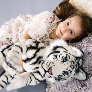 May include: A young girl in a white floral dress is lying on a pillow with a white tiger plush toy. The pillow is made of a soft, ruffled fabric.