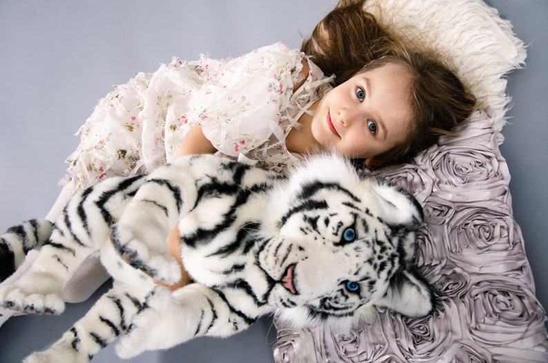 May include: A young girl in a white floral dress is lying on a pillow with a white tiger plush toy. The pillow is made of a soft, ruffled fabric.