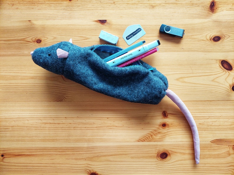 May include: A gray felt pencil case shaped like a mouse with a pink tail. The case is open and filled with blue and pink pens, a blue eraser, and a silver USB drive.