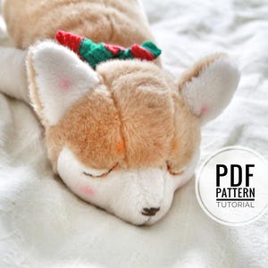 Peut inclure: Un chien en peluche avec une fourrure brune et des marques blanches est allongé sur une couverture blanche. Le chien porte un nœud vert et rouge autour du cou. Le texte "PDF PATTERN TUTORIAL" est visible dans un cercle blanc.