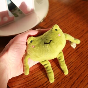 May include: A green plush frog toy with a smiling face and black eyes. The frog is being held in a hand.