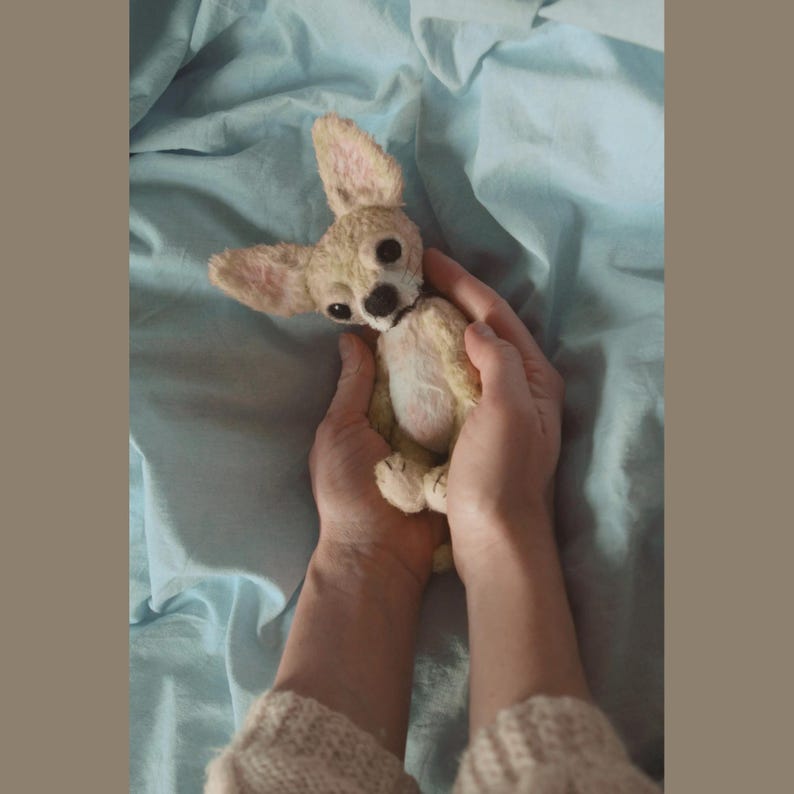 May include: A small, light brown and white plush chihuahua dog toy. The toy is being held in the hands of a person.