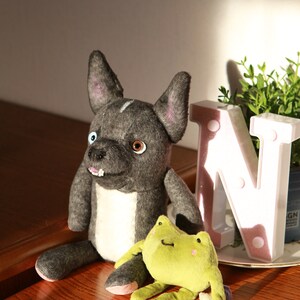May include: A gray and white plush dog toy with a black nose and brown eyes. The dog toy is sitting on a wooden surface next to a green plush frog toy. The frog toy has a white belly and pink cheeks.
