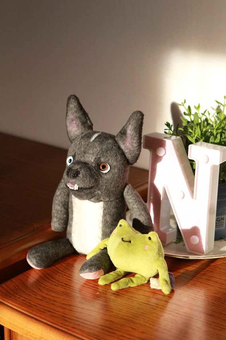 May include: A gray and white plush dog toy with a black nose and brown eyes. The dog toy is sitting on a wooden surface next to a green plush frog toy. The frog toy has a white belly and pink cheeks.