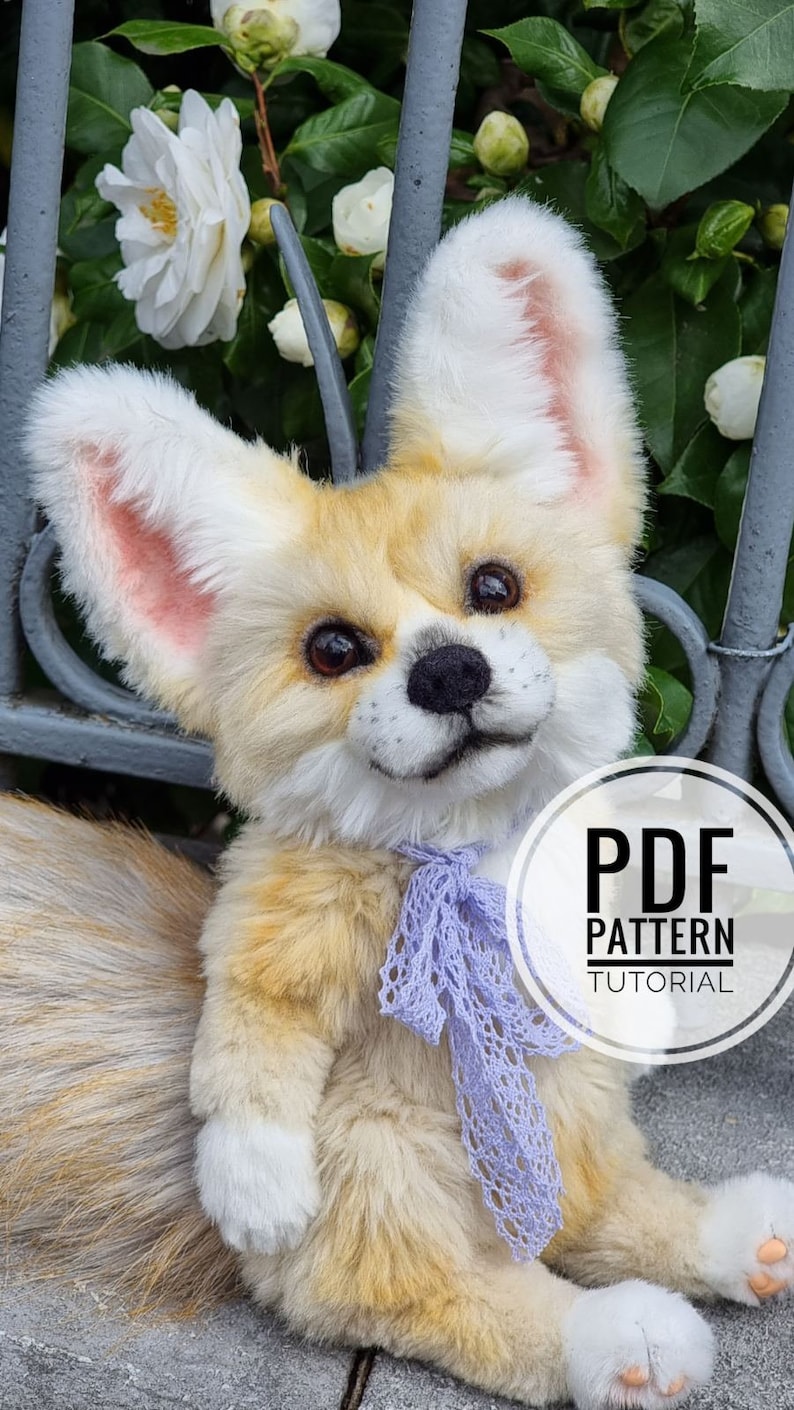 May include: A plush toy fox with a light brown and white fur, wearing a purple ribbon. The toy is sitting on a gray surface with a white flower in the background. The text "PDF PATTERN TUTORIAL" is on a white circle.