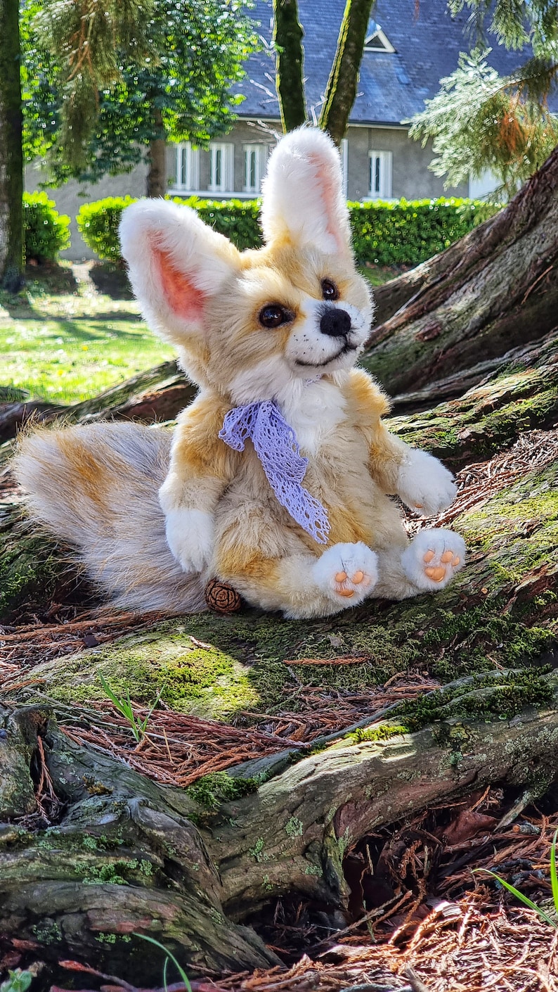 May include: A plush toy fox with a long, bushy tail. The fox is sitting on a tree root and has a purple ribbon around its neck. The fox has a light brown and white fur with a pink nose and paws.