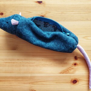 May include: A blue fabric pencil case shaped like a mouse with a pink tail and a blue zipper. The case is open and the inside is lined with a blue and white fabric with a small mouse print.