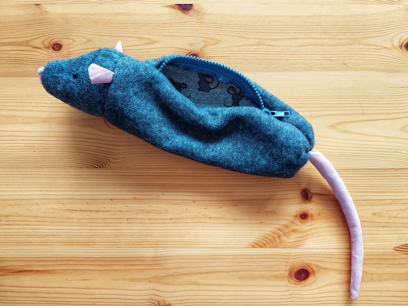 May include: A blue fabric pencil case shaped like a mouse with a pink tail and a blue zipper. The case is open and the inside is lined with a blue and white fabric with a small mouse print.