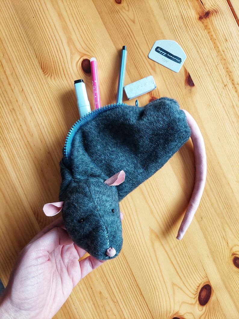 May include: A gray felt pencil case shaped like a mouse with pink ears and a tail. The case is open and contains a pink highlighter, a blue highlighter, a black marker, a blue pen, and two white erasers. One eraser has the text "Happy memories" written on it.