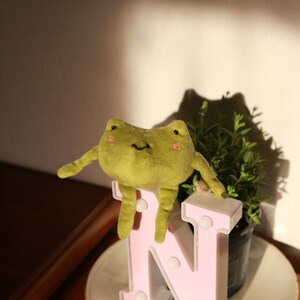 May include: A plush green frog toy sitting on a pink letter N with white lights. The letter is on a white saucer with a floral design.