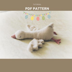 Op de afbeelding: Een wit pluche gansenspeelgoed met roze accenten ligt op een wit beddengoed. De gans heeft een lange nek en een kleine snavel. Een string met kleurrijke paaseieren vlaggen hangt boven de gans.