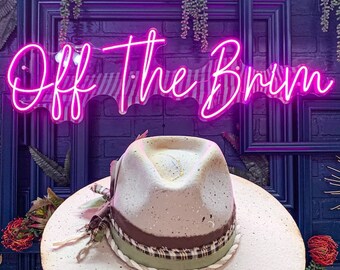May include: A light gray felt hat with a brown and green ribbon and a white rope trim. The hat is sitting on a white surface in front of a pink neon sign that says "Off the Brim".