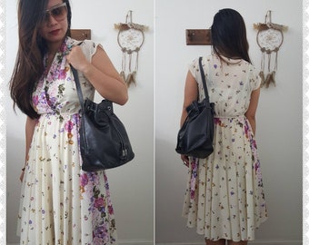 May include: A cream-coloured midi dress featuring a vibrant floral print with purple, pink, and green flowers, a V-neckline, and short sleeves. The dress is paired with a black leather bucket bag that has a drawstring closure and a shoulder strap.