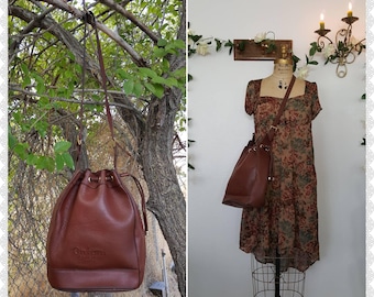 May include: A brown leather bucket bag with a drawstring closure, gold-tone metal hardware, and a long adjustable shoulder strap. The front of the bag features the embossed text Galani. The bag is shown hanging from a tree and on a dress form.