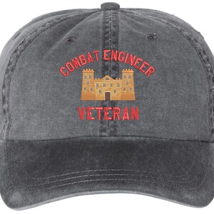May include: A charcoal gray baseball cap with the words "COMBAT ENGINEER VETERAN" embroidered in red. A tan and brown castle graphic is centered between the text. The cap has a curved brim and a fabric strap closure.