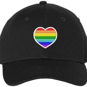 May include: Black baseball cap with a rainbow heart patch. The heart features horizontal stripes in red, orange, yellow, green, blue, and purple. The cap has a curved brim and a button on top.