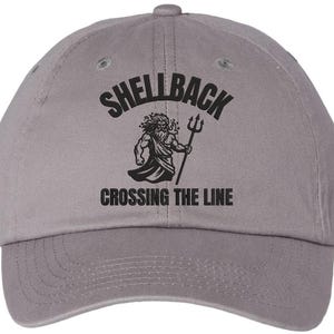 May include: A gray baseball cap with the word "SHELLBACK" arched across the top. Below the text is a black illustration of a muscular figure holding a trident. The text "CROSSING THE LINE" is below the illustration.