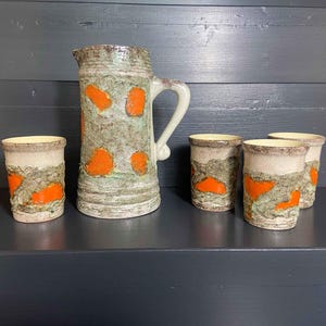 Peut inclure: Un pichet en céramique et quatre gobelets assortis. L'ensemble présente un glaçage gris et vert moucheté avec de grands accents orange. Le pichet a une anse et un bec. Les gobelets sont cylindriques avec un design similaire.