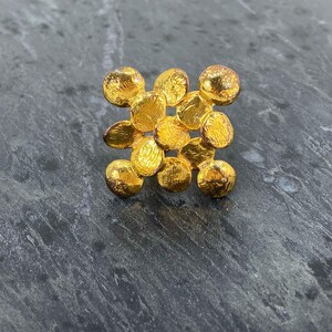 Op de afbeelding: Een goudkleurige ring met een vierkant cluster van negen kleine, ronde, getextureerde schijfjes in een rasterpatroon. Het oppervlak van elke schijf heeft een gehamerde, onregelmatige afwerking die licht reflecteert voor een heldere, metalen uitstraling.