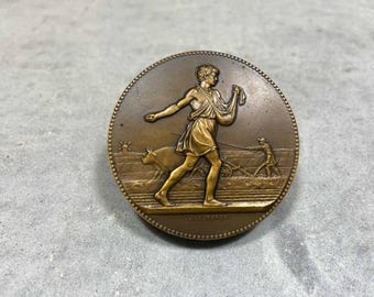 Puede incluir: Una medalla de bronce que representa a un hombre arando un campo con una vaca. La medalla tiene un diseño en relieve y está rodeada por un borde de cuentas. El texto "VALLORANCE" está inscrito en la parte inferior de la medalla.