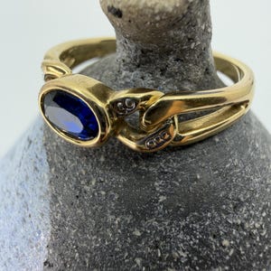 Può includere: Un anello in oro con una pietra preziosa blu scuro dal taglio ovale incastonata in una lunetta. La fascia presenta un design a maglie di catena con piccoli accenti color argento incastonati. Il metallo ha una finitura lucida e l'anello è appoggiato su un oggetto testurizzato.