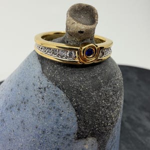 May include: A gold band ring featuring a central circular blue stone setting flanked by a channel of small clear stones. The ring has a polished gold finish and a structured design with a raised section around the blue stone, creating a distinct profile.