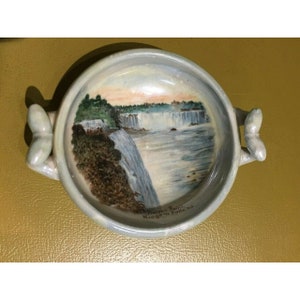 May include: A small, white ceramic dish with two butterfly-shaped handles. The dish features a painted image of Niagara Falls with the text "Prospect Point Niagara Falls, N.Y."