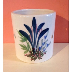 Pode incluir: Casti&ccedil;al de cer&acirc;mica branca com um design floral pintado &agrave; m&atilde;o. O design apresenta elementos bot&acirc;nicos azuis, verdes e rosa. O casti&ccedil;al tem dois pequenos orif&iacute;cios e &eacute; de formato cil&iacute;ndrico.