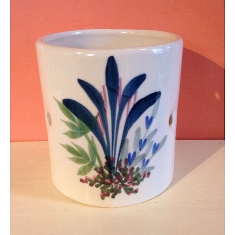 Pode incluir: Casti&ccedil;al de cer&acirc;mica branca com um design floral pintado &agrave; m&atilde;o. O design apresenta elementos bot&acirc;nicos azuis, verdes e rosa. O casti&ccedil;al tem dois pequenos orif&iacute;cios e &eacute; de formato cil&iacute;ndrico.