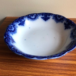 May include: A white ceramic bowl with a scalloped edge and a blue floral design around the rim. The bowl has a simple, elegant design, perfect for serving food or as a decorative piece. The bowl is set on a wooden surface.