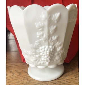 May include: White milk glass vase with a grapevine design. The vase has a scalloped rim and a pedestal base.