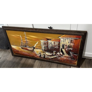 May include: A framed painting of a seascape with a sailboat docked at a waterfront village. The painting is done in a stylized, abstract style with bold colors and brushstrokes. The colors are mostly warm tones of yellow, orange, and brown.