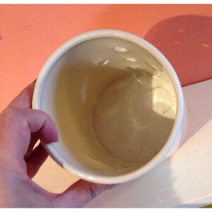 Pode incluir: Um vaso de cer&acirc;mica branco com formato arredondado e uma s&eacute;rie de pequenos orif&iacute;cios perto da borda superior. O interior &eacute; bege claro. O vaso &eacute; segurado por uma m&atilde;o, com uma parede cor de p&ecirc;ssego ao fundo.
