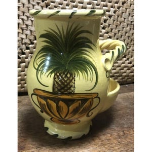 May include: A yellow ceramic pitcher with a twisted handle and a painted design of a pineapple plant in a pot. The pitcher has a small spout on the side.