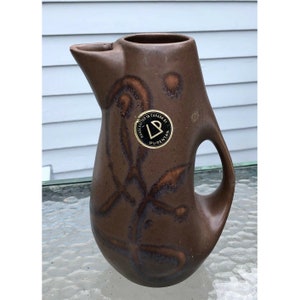 May include: A brown ceramic pitcher with a unique, abstract design. It has a wide, rounded base and a narrow spout. The pitcher has a handle on the side and a label that reads "Handcrafted in Canada by Purensan".