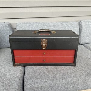 May include: A black and red metal toolbox with two drawers. The toolbox has a metal latch and a handle.