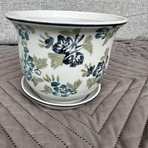 May include: White ceramic planter with a blue and green floral design. The planter has a matching saucer.