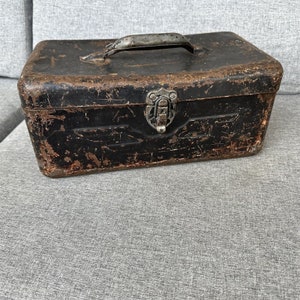 May include: A vintage metal toolbox with a worn black finish and a silver latch. The toolbox has a metal handle on the top.