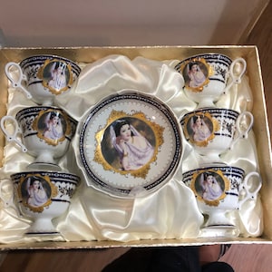May include: A set of six white porcelain teacups and saucers with gold trim and a blue and gold floral pattern. Each cup features a portrait of a woman in a white dress with a gold crown.