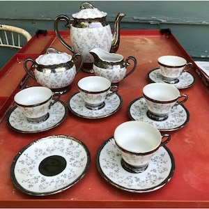 May include: A vintage silver and white tea set with a teapot, creamer, sugar bowl, and six cups and saucers. The set features a delicate floral pattern.