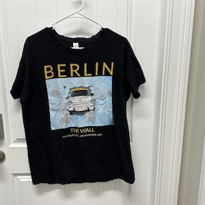 May include: Black t-shirt with the word "BERLIN" in gold letters. The shirt features an image of a car breaking through a wall, with the text "THE WALL 13. AUGUST 1961 - 09. NOVEMBER 1989". The shirt is hanging on a white hanger.