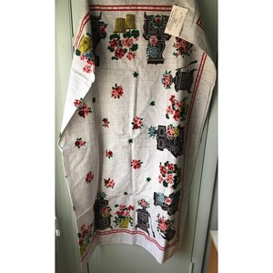 May include: A vintage white kitchen towel with a repeating pattern of floral arrangements and black stoves. The towel has a red border and is made of a linen-like fabric. A tag is attached to the top.