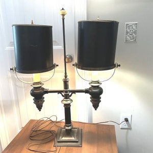 May include: A two-light antique brass table lamp with black lampshades. The lamp has a decorative design with a central column and two arms extending out to the lampshades. The lampshades are cylindrical and have a black finish.