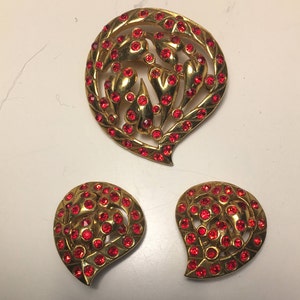 May include: A gold-tone brooch and earrings set with red rhinestone accents. The brooch features a stylized floral design and the earrings are heart-shaped.