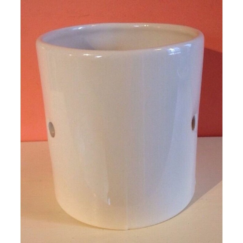 Pode incluir: Um recipiente cil&iacute;ndrico de cer&acirc;mica branca com dois pequenos orif&iacute;cios espa&ccedil;ados. O recipiente tem um acabamento liso e brilhante e uma forma ligeiramente curva. O fundo &eacute; de cor laranja quente.