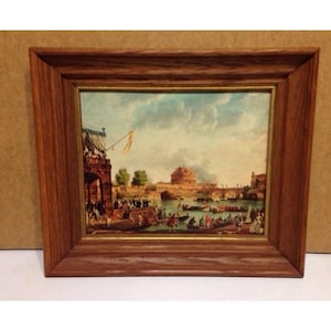May include: A framed print of a painting depicting a scene of a city with a bridge and boats on a river. The painting is in a brown wooden frame with a gold trim.