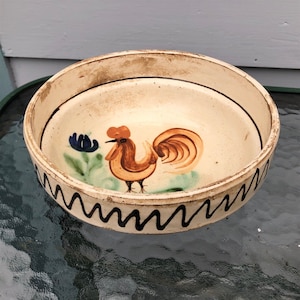 May include: A white ceramic bowl with a hand-painted rooster design. The rooster is brown with a red comb and wattle, and has a green and blue flower behind it. The bowl has a black wavy line around the rim.