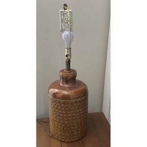 May include: A brown ceramic table lamp with a woven pattern. The lamp has a gold-colored base and a white light bulb.