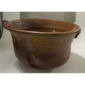 May include: A large, brown ceramic bowl with two small handles on the sides. The bowl has a rough, textured surface and a slightly uneven rim.